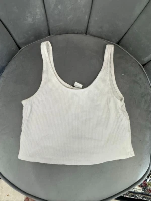 Vit ribbad croppad linnetopp - Säljer en enkel och fräsch vit croppad linnetopp med ribbad struktur. Toppen är ärmlös och har breda axelband, perfekt för varma dagar eller som layering-piece. Passar till det mesta och är ett måste i garderoben!