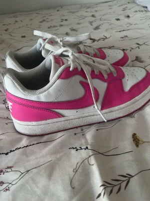  Nike skor  - Nike sneakers i vitt och starkt rosa med klassisk swoosh på sidan. Skorna har vita snören, perforerad tåbox och lågt skaft. Tillverkade i läder och textil med en vit och rosa yttersula. Perfekta för dig som vill sticka ut med färg.