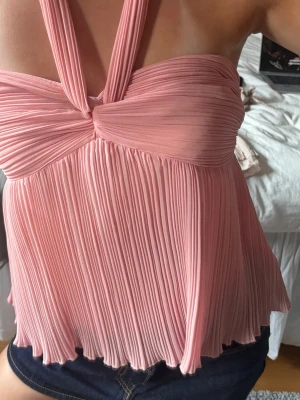 Plisserad rosa halterneck topp - Säljer en superfin rosa halterneck topp med plisserat tyg och twistad detalj framtill. Toppen är ärmlös och har en luftig, flowy passform som ger en riktigt söt och trendig look. Perfekt till sommaren och enkel att styla med jeans eller kjol.