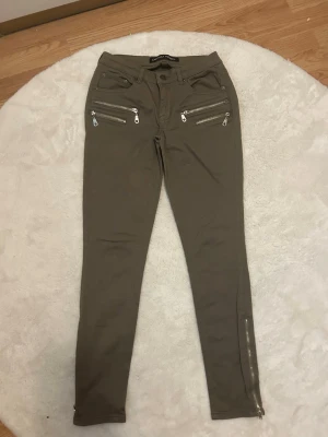 Olivgröna skinny jeans med dragkedjor - Snygga olivgröna skinny jeans med flera silverfärgade dragkedjor framtill och vid benslut. Jeansen har medellång midja och klassisk femficksdesign. Materialet är stretchigt för en bekväm passform och ger en cool, modern look.