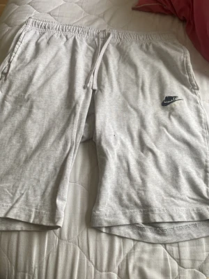 Ljusgrå shorts från Nike med snörning - Snygga ljusgrå shorts från Nike med elastisk midja och snörning. Perfekta för sommaren eller träning. Mjukt material och klassisk Nike-logga på ena benet. Avslappnad passform och bekväma fickor på sidorna.