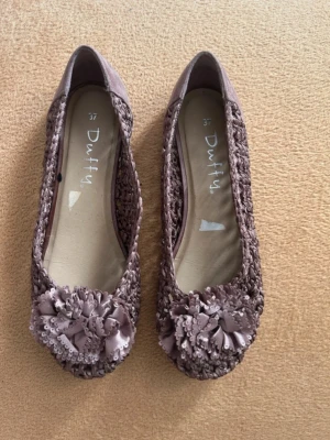 Rosa/lila ballerinaskor med blommor - Säljer ett par rosa/lila ballerinaskor från Duffy i storlek 37. Skorna har en flätad struktur och dekoreras med stora blommor framtill. Materialet är textil med detaljer i satinliknande tyg. Perfekta för dig som gillar romantisk och söt stil.