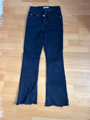 Svarta bootcut jeans med rå kant - Säljer ett par svarta bootcut jeans med hög midja och rå, fransad kant nedtill. Jeansen har klassisk femficksdesign och knappgylf. Perfekta för dig som gillar en avslappnad men trendig stil.