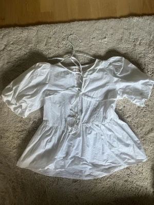 Vit puffärmad blus från Nelly - Söt vit blus från Nelly med puffiga korta ärmar och knytning framtill. Blusen har en lätt utsvängd passform och är gjord i ett tunt, krispigt tyg som ger en luftig känsla. Perfekt för en trendig och avslappnad look.
