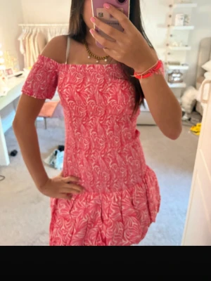 Rosa offshoulder kortklänning - Söt rosa kortklänning med vitt blommönster och offshoulder-ärmar. Klänningen har volang nedtill och en figurnära passform som ger en feminin siluett. Perfekt för varma dagar och somriga tillfällen.