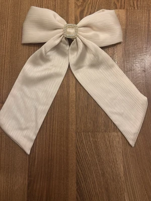 Stor cremefärgad hårrosett från Glitter  - Helt oanvänd cremefärgad hårrosett i ribbat tyg med lång knytning och dekorativt spänne med pärlor och strass i mitten. Fästs enkelt med metallklämma på baksidan. Perfekt för att ge en extra touch till din frisyr. Obs säljer endast via plicks köpknapp. 