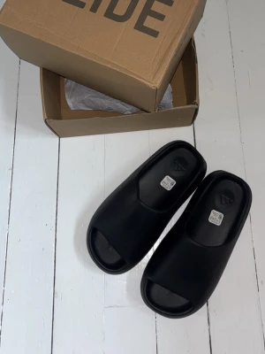 Yeezy slides tofflor svarta - Säljer ett par svarta adidas yeezy slides tofflor med öppen tå och ribbad sula. Tillverkade i ett mjukt och flexibelt syntetmaterial som ger skön komfort. Perfekta för att glida runt hemma eller på stranden. Enkel och stilren design. Storlek 45