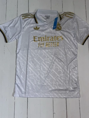  Real Madrid Tröja Adidas - Snygg vit Real Madrid matchtröja från Adidas med gulddetaljer och klubbmärke på bröstet. Tröjan har korta ärmar, krage och diskret mönster i tyget. Tillverkad i lätt och ventilerande material, perfekt för fotboll eller träning. Storlek L