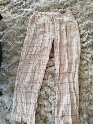 Rutiga pyjamasbyxor i beige och vinrött - Mysiga pyjamasbyxor i mjukt tyg med rosa bas och rutigt mönster i olika rosa nyanser. Byxorna har resår i midjan och sidofickor, perfekta för chillkvällar hemma. Klassisk rak passform och skön känsla mot huden.