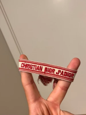 Christian Dior J'Adior armband - Snyggt armband från Christian Dior med texten 'J'Adior' i rött på en beige bakgrund. Armbandet är vävt i tyg och har röda fransar i ändarna. Perfekt accessoar för att lyfta din outfit med en lyxig touch.