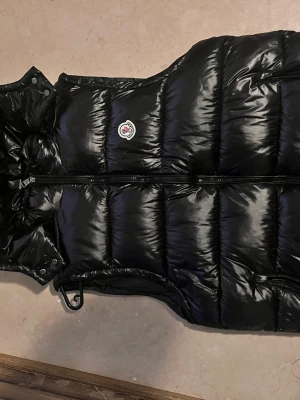 Svart dunväst med huva från Moncler - Snygg svart dunväst från Moncler med glansig finish och vadderad design. Västen har dragkedja framtill, två sidofickor med dragkedja och en hög krage med avtagbar huva. Ikonisk Moncler-logga på bröstet. Perfekt för lager-på-lager under kyliga dagar.