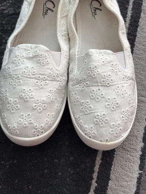 Vita ballerinaskor med broderade blommor - Säljer ett par vita ballerinaskor med söt broderad spets och små blommor över hela skon. Skorna har elastiska partier vid öppningen för extra komfort och en vit sula. Perfekta för dig som gillar en romantisk och feminin stil.