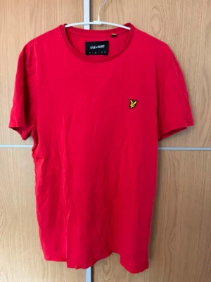 Röd t-shirt från Lyle & Scott - Snygg röd t-shirt från Lyle & Scott med klassisk gul logga på bröstet. T-shirten har rund halsringning och korta ärmar. Tillverkad i mjuk bomull som känns skön mot huden. Perfekt för en avslappnad och stilren look.