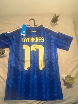 Blå gyökeres Sverige fotbollströja Adidas - Säljer en blå Sverige fotbollströja från Adidas med gult tryck, nummer 17 och namnet Gyökeres på ryggen. Tröjan har korta ärmar, gula detaljer och ett mönster i blå nyanser. Perfekt för dig som vill stötta landslaget på match!