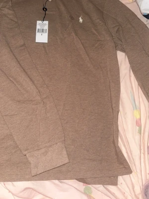 Brun långärmad henley från Polo Ralph Lauren - Säljer en brun långärmad henley från Polo Ralph Lauren med klassisk logga broderad på bröstet. Skjortan har knappar vid halsen och är gjord i mjuk bomull, perfekt för en avslappnad och stilren look.