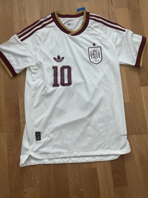 Vit Spanien tröja Adidas nr 10 lamine Yamal - Säljer en vit Spanien fotbollströja från Adidas med vinröda detaljer och nummer 10. Tröjan har LAMINE YAMAL tryckt på ryggen, guldiga muddar och klassiska Adidas-ränder på axlarna. Perfekt för dig som älskar fotboll och vill sticka ut på planen.