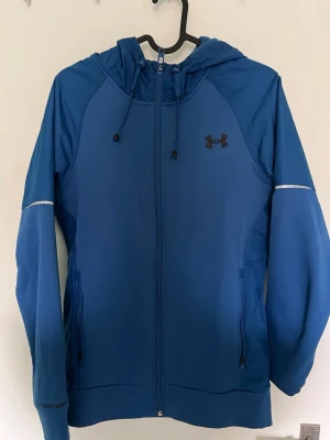 Blå hoodie från Under Armour - Snygg blå hoodie från Under Armour med dragkedja hela vägen, justerbar huva och två fickor framtill. Tillverkad i ett mjukt och sportigt material, perfekt för träning eller chill. Logga på bröstet och text på ryggen ger en clean look. Inga slitningar osv. Hör av er om intresserad
