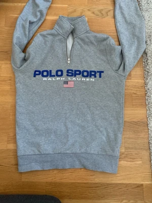 Grå halvzip från Ralph Lauren - Snygg grå sweatshirt med halvzip och hög krage från Polo Sport Ralph Lauren. Stor blå logga och amerikansk flagga framtill. Tillverkad i mjukt sweatshirtmaterial, perfekt för en sportig och avslappnad stil.