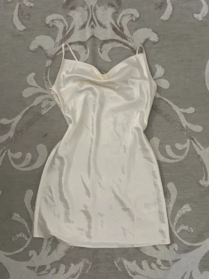 Vit kort satinklänning med smala band - Säljer en elegant vit kort klänning i satin med smala axelband och mjukt draperad urringning. Perfekt för dig som gillar en stilren och chic look. Klänningen har en figurnära passform och glänsande material som ger en lyxig känsla.