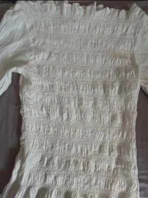 Vit offshoulder klänning Stories S - Supersöt vit offshoulder klänning från Stories i storlek S. Klänningen har smock över hela kroppen och volangdetalj upptill. Perfekt för dig som gillar romantisk och trendig stil.