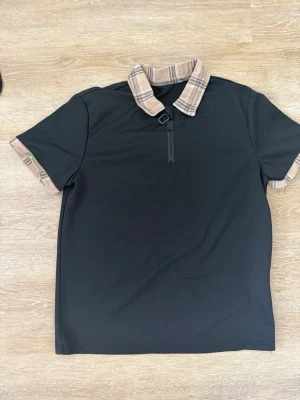 Svart t-shirt med rutiga detaljer - Svart t-shirt med korta ärmar och rutigt mönster på krage och ärmslut. Tröjan har en half zip-dragkedja framtill och en loose passform. Perfekt för dig som vill ha en unik twist på din vardagsstil.