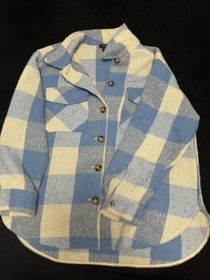 Blåvit rutig overshirt från Lindex - Kofta från Lindex i blåvitt rutigt mönster. Modellen har stora bröstfickor, knappar framtill och en klassisk krage. Materialet är grovt vävt och ger en mysig och trendig känsla. Perfekt för lager på lager och en chill stil.