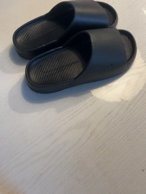 Svarta nike tofflor  - Säljer ett par svarta slides sandaler i syntetmaterial med ribbad innersula och bred rem över foten. Minimalistisk design och bekväm passform, perfekta för chill dagar eller att glida runt hemma.