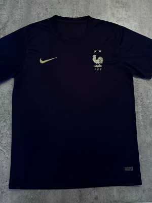 Blå Nike Frankrike fotbollströja - Mörkblå fotbollströja från Nike med Frankrikes landslagsemblem och två stjärnor broderade på bröstet. Tröjan har rund hals, korta ärmar och är tillverkad i ett lätt, ventilerande material som passar perfekt till träning eller match.