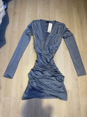 Grå omlottklänning med lång ärm - Snygg grå kortklänning med omlott och djup v-ringning. Klänningen har långa ärmar och är figurnära med draperad effekt framtill. Perfekt för dig som gillar en stilren och trendig look.