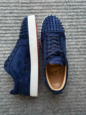 Mörkblå sneakers med nitar - Snygga mörkblå sneakers med matta nitar över hela tån och ovandelen. Skorna har vit sula med röd undersida och är gjorda i mockaliknande material. Klassisk snörning och låg profil gör dem till ett statement i varje outfit.