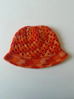 Orange virkad bucket hat - Handgjord bucket hat i virkad bomull, i orangea nyanser.  Hatten har ett luftigt mönster och brett brätte, perfekta för en trendig och avslappnad stil. Säljs styckvis. 