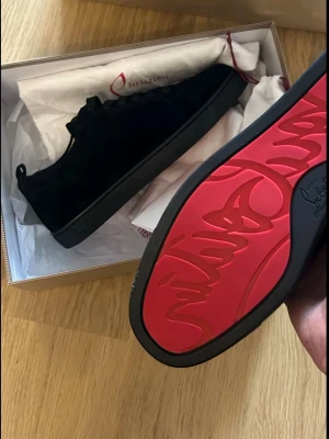 Christian Louboutin - Stilrena svarta sneakers i mocka från Christian Louboutin med ikonisk röd sula och diskret logga undertill. Klassisk låg modell med svarta skosnören och minimalistisk design. Perfekta för dig som vill ha en lyxig och trendig look.