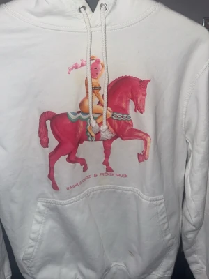 Vit hoodie med tryck av rosa häst - Vit hoodie med stor färgglad print framtill som visar en person med rosa mask ridandes på en rosa häst. Texten 'Rasmus Gozzi & Fröken Snusk' under motivet. Klassisk känguruficka och dragsko i huvan. Mjuk bomullskänsla och avslappnad passform.