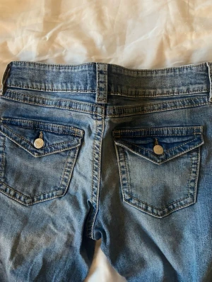 Blå bootcut jeans  - Säljer ett par blå bootcut jeans med lätt slitna detaljer och klassisk femficksmodell. Jeansen har en snygg tvätt och är tillverkade i denim med lite stretch för extra komfort. Perfekta för en avslappnad och trendig look.