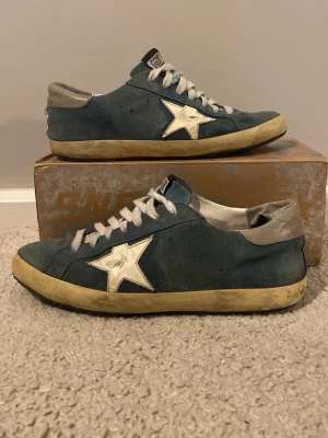 Blå GGDB sneakers med vit stjärna - Snygga blå sneakers från Golden Goose med vit stjärndetalj på sidan och grå häl. Skorna har vita snören, beige sula och GGDB-logga på plösen. Ovandelen är i mocka och designen har en vintagekänsla som sticker ut. En väldigt bra skomakare har lagat sulan under och byt ut hrla sulan mot en ny vid pris 1500 bara för sil byte