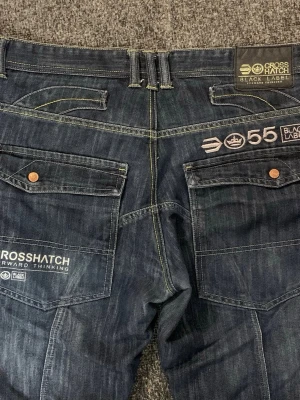 Mörkblå jeans från Crosshatch - Snygga mörkblå jeans från Crosshatch Black Label med kontrastsömmar och flera detaljerade broderier och tryck på bakfickorna. Klassisk femficksmodell med knappgylf och bälteshällor. Jeansen har en modern passform och robust denimkänsla. Custom