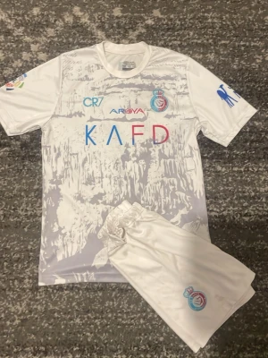 Vit fotbollströja och shorts set - Säljer ett vitt fotbollskit med tröja och shorts. Tröjan har tryck i blått och rött, CR7-logga, klubbmärke och flera sponsorer på bröstet och ärmarna. Materialet är lätt och andas, perfekt för träning eller match. Matchande vita shorts med klubbmärke ingår. storlek 152, skicket är bra på tröjan men mindre bra på shortsen(syns på bilderna) men fläckarna på tröjan syns ej på 