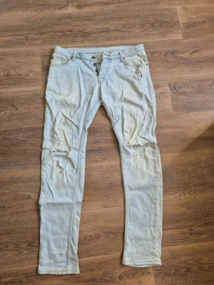 Ljusblå slitna jeans från Hammond - Säljer ett par ljusblå jeans från Hammond med skinny fit och slitningar på både lår och knän. Jeansen har klassisk femficksmodell, bälteshällor och knappgylf. Perfekta för dig som gillar en avslappnad och trendig stil. Modell: Nevada
