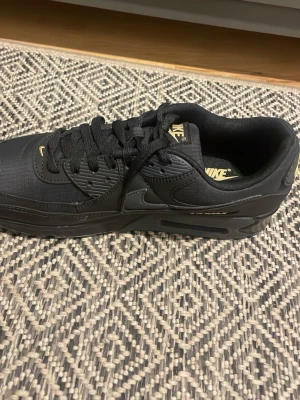 Nike Air Max svarta sneakers - Snygga svarta Nike Air Max sneakers med gulddetaljer på loggan och hälen. Skorna har en klassisk låg siluett, vadderad krage och synlig Air-dämpning i sulan. Ovandelen är i textil och syntet för en modern look. Perfekta för dig som gillar streetwear och sportig stil.