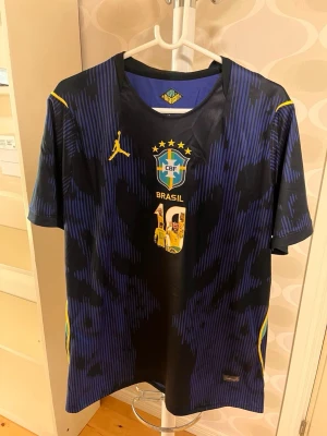 Brasilien Neymar Jr matchtröja blå - Snygg blå och svart matchtröja för Brasilien med Neymar Jr:s namn och nummer 10 på ryggen. Tröjan har gula detaljer, CBF-logga och tryck med Neymar-bilder. Perfekt för fotbollsfans som vill sticka ut på planen eller läktaren. passar som en XL