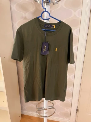 Olivgrön t-shirt från Polo Ralph Lauren - Snygg olivgrön t-shirt från Polo Ralph Lauren med klassisk gul logga broderad på bröstet. T-shirten har rund halsringning och korta ärmar. Tillverkad i mjuk bomull för en bekväm känsla. Perfekt till jeans eller shorts.