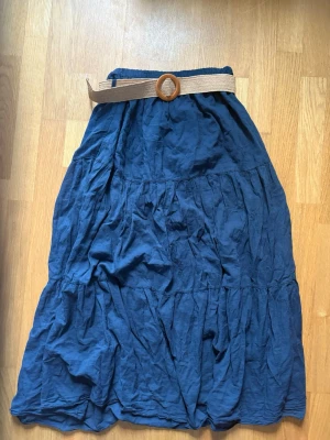 Marinblå långkjol med bälte - Säljer en marinblå långkjol i bomull med beige bälte och rund spänne. Kjolen har en avslappnad passform och är perfekt för varma dagar. Materialet känns lätt och luftigt, och kjolen har en snygg, volangig struktur. Den är köpt i Italien, därför har den ingen prislapp eller tydlig storlek men den passar mig perfekt som är 169cm lång och har storlek S. 