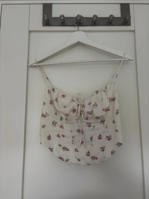 Blommig linnetopp med snörning - Söt ljus linnetopp med tunna axelband och små rosa blommor. Toppen har snörning både fram och bak för en justerbar passform och en feminin look. Perfekt för varma dagar och somriga outfits.