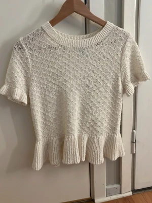 H&M topp - Söt vit stickad topp från H&M Divided med korta ärmar och volangkant nedtill. Tröjan har rund halsringning och ett diskret strukturmönster i stickningen. Perfekt till jeans eller kjol för en fräsch look.