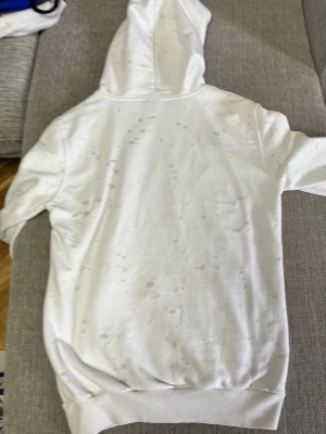 Vit hoodie från Givenchy Paris - Säljer en vit hoodie från Givenchy Paris med svart logotyptryck på bröstet. Hoodien har huva med metallöljetter och ett distressed mönster över hela tyget för en trendig look. Tillverkad i mjukt bomullsmaterial och har ribbade muddar vid ärmslut och nederkant.