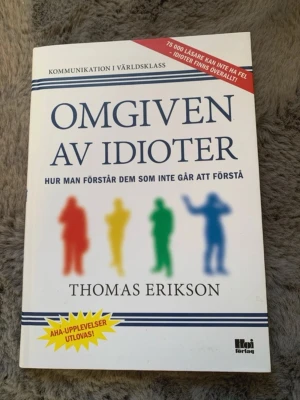 Omgiven av idioter av Thomas Erikson - Boken 'Omgiven av idioter' av Thomas Erikson handlar om kommunikation och hur man förstår olika personlighetstyper. Omslaget är vitt med färgglada siluetter i rött, gult, grönt och blått. Utgiven av Hoi förlag. Perfekt för dig som vill förstå människor bättre.