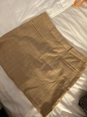 Beige rutig kortkjol - Snygg beige kortkjol med rutigt mönster och diskreta fickor fram. Kjolen har bälteshällor och klassisk rak passform, perfekt till en trendig vardagslook. Materialet känns mjukt och lätt, troligen bomull eller polyester.