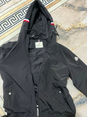 Svart vindjacka från Moncler - Snygg svart vindjacka från Moncler med huva och dragkedja framtill. Jackan har vita och röda detaljer på huvan, två fickor med tryckknappar och Moncler-logga på ärmen. Tillverkad i lätt och tåligt material, perfekt för blåsiga dagar.