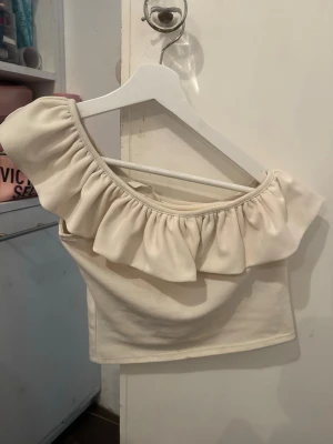 Vit omlottopp med volangdetalj - Säljer en söt vit topp med omlottkänsla och stor volang längs den ena axeln. Toppen är croppad och har en ärmlös design med en axel bar. Perfekt för dig som gillar trendiga och feminina plagg. Två minifläckar som nog lägg går att få bort!☺️