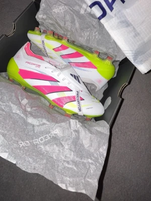 Adidas Predator Elite fotbollsskor vit/rosa - Säljer ett par Adidas Predator Elite fotbollsskor i vitt med rosa och neongula detaljer. De har använts ca. 10 gånger, men är i nyskick och relativt rena. Skorna har klassisk snörning, mönstrad sula för bra grepp och en sportig design med tydliga loggor. Perfekta för dig som vill sticka ut på planen. Påsen på bilden till skorna kommer även med. 
